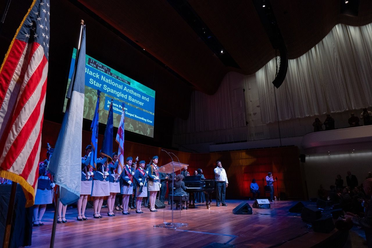 A group of uniformed individuals holding flags stands on a stage next to a lectern. One person is singing near a piano, while others look on. A large screen in the background displays text about the Black National Anthem and Star Spangled Banner performance. The scene is set in a spacious auditorium with an audience.